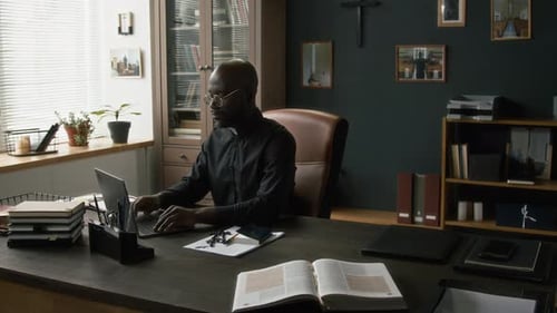 Focused Young Priest Typing on Laptop while Working at Large Table in His Office