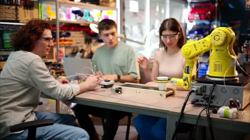 Young happy engineers programming an yellow robotic arm in the workshop to grab cardboard water glas