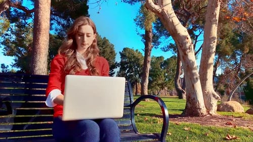 A happy, attractive, young, caucasian woman at the park, sitting on a bench, typing on her laptop. S