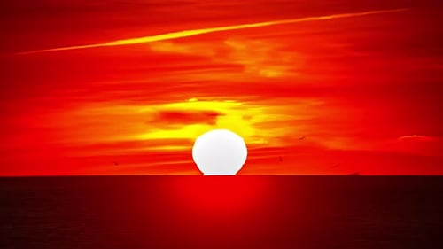 Round sun orb in vivid red sunset sky dips below horizon over ocean, timelapse