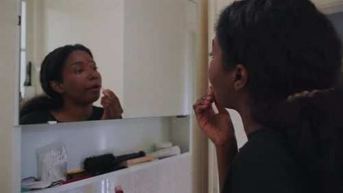 African American woman applying lip balm while looking at her reflection in bathroom mirror