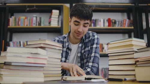 Young Man Studying at a Library Surrounded By Stacks of Books While Focusing on His Notes During the
