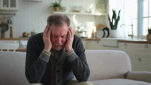 Senior Man Massages Temples on Couch at Home