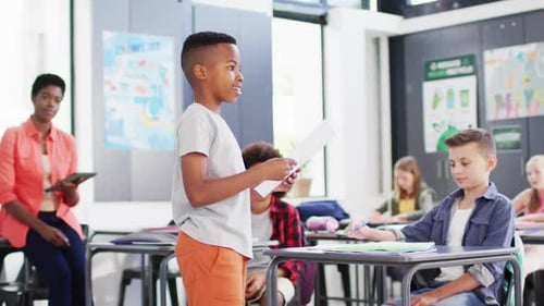 Diverse female teacher and happy schoolchildren at desks reciting in school classroom