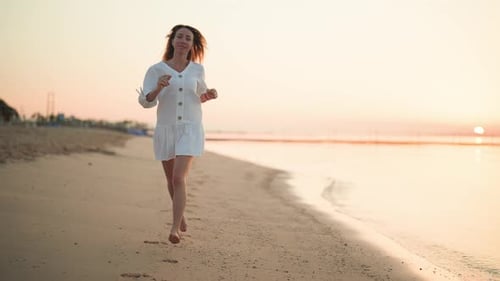 Entspannte blonde Touristin in luftigem weißem Kleid, die bei Sonnenuntergang am Meer entlang läuft, Sandstrand, weiblich