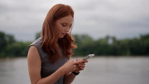 Closeup of Cute Longhaired Young Woman Seen Standing By River Using Smartphone Checking Emails or