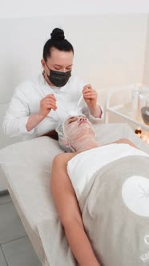 Esteticista de terapia enzimática aplicando creme médico hidratante no rosto de uma jovem adulta com pincéis