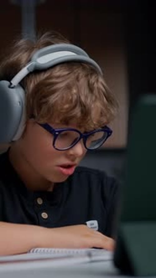 Young Boy with Curly Hair and Blue Glasses Wearing Large Headphones Intently Using a Tablet and