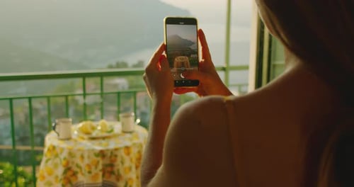 A woman on a balcony uses her phone to photograph the scenic view of the Amalfi Coast, with a breakf