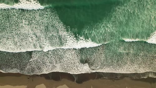 Foamy ocean waves rolling to tropical shore with dark sand, aerial top down view