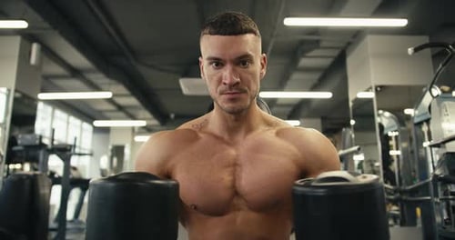 Porträt eines jungen Bodybuilders mit nacktem Torso, der in die Kamera schaut und schwarze Hanteln hält