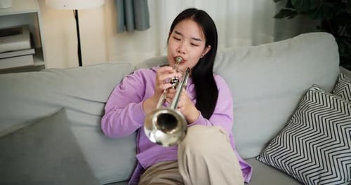 A young woman practices playing the trumpet while sitting on a sofa in a cozy living room.