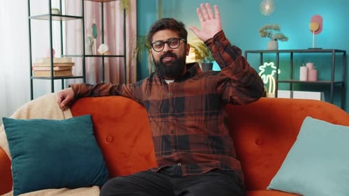 Smiling Man Waving Hello from Orange Couch Indoors
