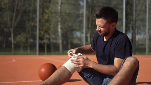 Man Sit on Basketball Court and Bandages Knee with Elastic Bandage After Injury
