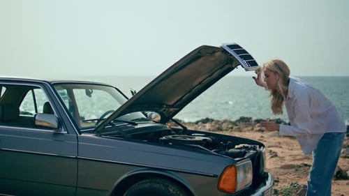 Confused young woman closing car hood at sunny ocean shore feeling disappointed