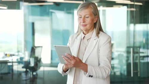 Confident senior gray haired businesswoman is using digital tablet standing in business office.