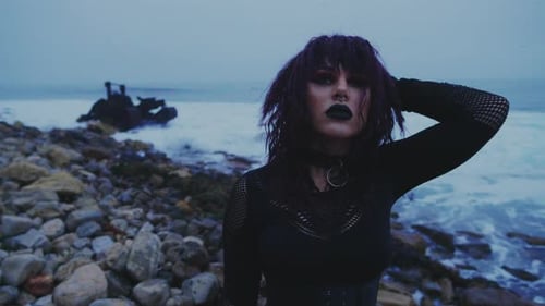 Una figura enmascarada vestida de negro se encuentra en la orilla del mar y se quita una máscara rota, revelando a una niña gótica