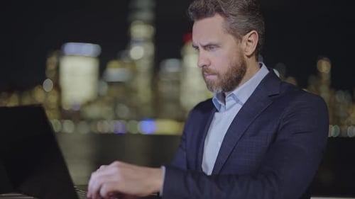 Successful Businessman Working on Laptop Computer in Big City Late in the Evening Working Overtime