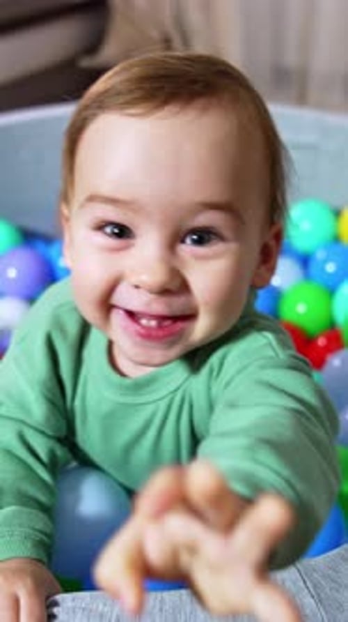 Joyful happy toddler smiling broadly stands in soft basin filled with colorful balls.