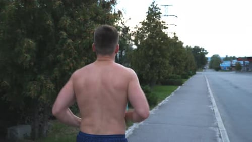 Rear View of Young Muscular Sportsman Jogging Along Sidewalk Near Forest at Summer Day