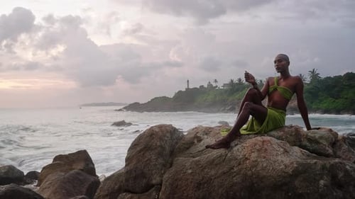 Divine Nonbinary Black Person in Long Open Dress Brass Jewelry Poses on Rocky Hill Top Above