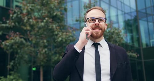 Un hombre de negocios con traje y elegantes anteojos se para en la calle de la ciudad mientras llama por teléfono inteligente.