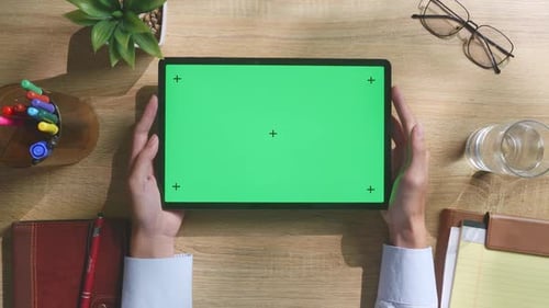 Top down view of man's hands holding a green screen tablet on a wooden desk