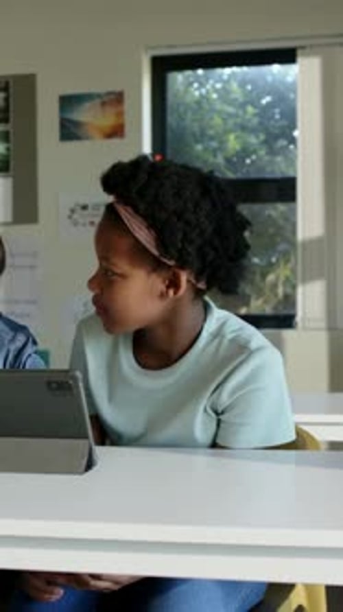 Vertical video: Noting peer, girl in mint top pink band turning left at classroom desk with tablet