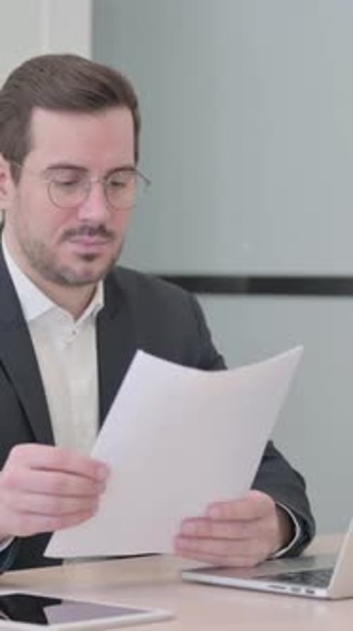 Man Reads Documents and Types on Laptop at Desk