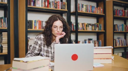 Étudiante aux cheveux bouclés étudiant dans une bibliothèque entourée de livres tout en utilisant un ordinateur portable pendant