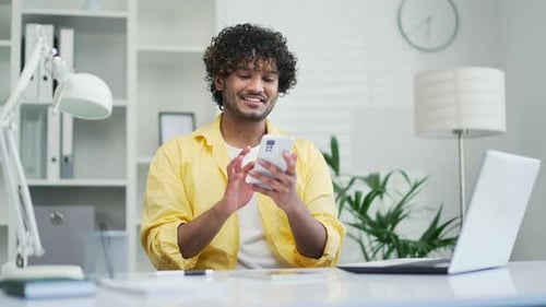 Happy handsome young man in yellow shirt uses phone in home office Mixed race student male typing br