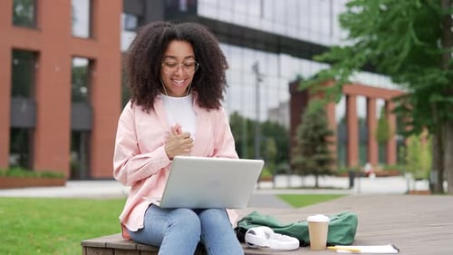 African american female student talking on video call using a laptop sitting on a bench in campus