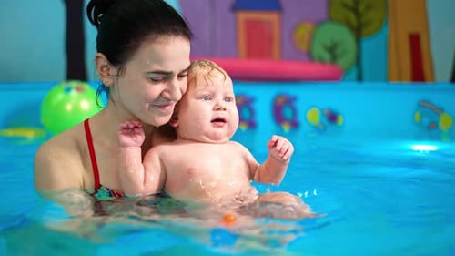Lovely cute Caucasian baby leaning on the shoulder of a woman in the swimming pool.