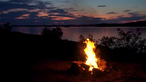 Campfire In The Beautiful Evening By The Lake With Mountains, Sunset And Clouds In The Background In