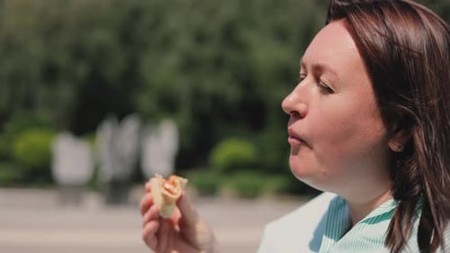 A brown-haired girl in a striped shirt sits on the wooden bench in the park and eats pizza