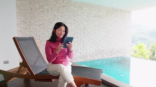 Asian woman relaxing on a pool bed and using digital tablet near the private pool during her morning
