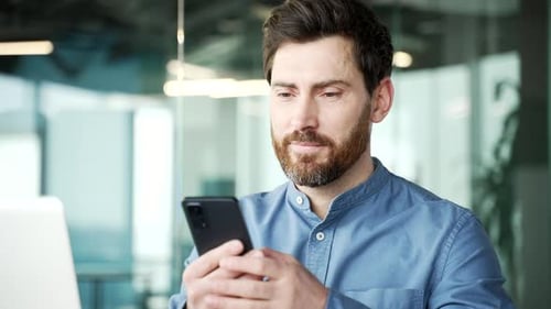 Happy bearded businessman is using phone while sitting in business office. Manager works on mobile