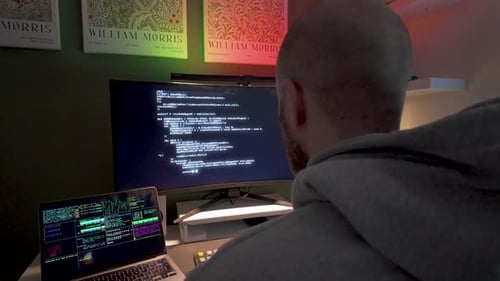 Back view of developer coding at dual monitors with colorful lights