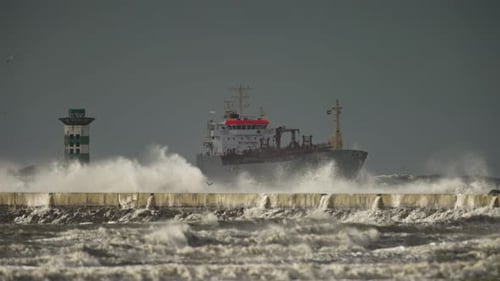 Storm waves crash over cement IJmuiden pier with ship in background, telephoto