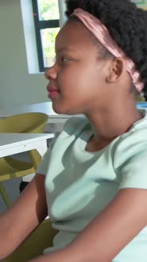 Vertical video: Turning student raising hand at school desk, answering, pink headband, copy space