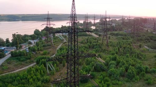 Structure of high-voltage lines outside the city at sunset. Trees and river. Camera motion back.