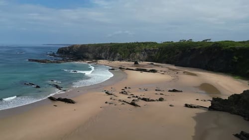 Aerial: Gentle waves wash sand beach below ocean coastline cliffs