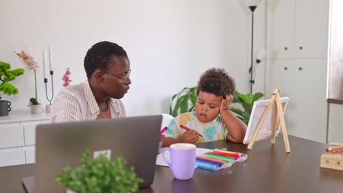 Working mother is talking to her toddler son who is drawing with colorful markers at the table