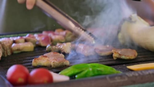 Tongs Turning Sizzling Meat Slices on a Smoky Barbecue Grill