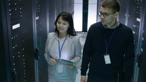 Caucasian Male and Asian Female IT Technicians Walking in Data Center with Rows of Rack Servers. Th