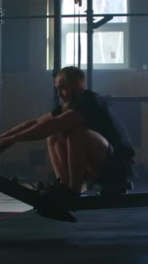 Focused Athletic Man Training on Indoor Rowing Machine in Dimly Lit Gym Demonstrating Powerful Back