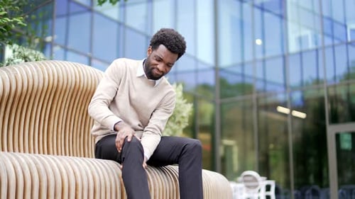 African american businessman suffers from painful severe knee joint pain sitting on bench on street