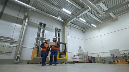 Engineers Stand in Terminal of Warehouse Looking in Laptop