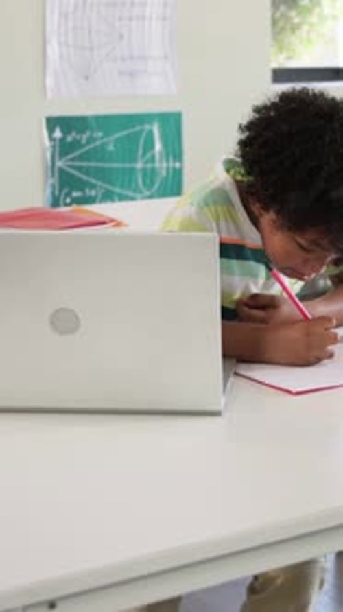 Vertical video: Writing kid in striped tee looking at laptop at desk, holding notes and pink pencil