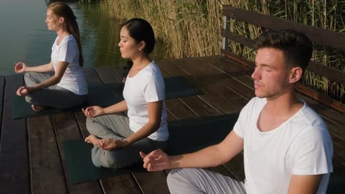 Group of people doing yoga exercises by the lake at sunset.